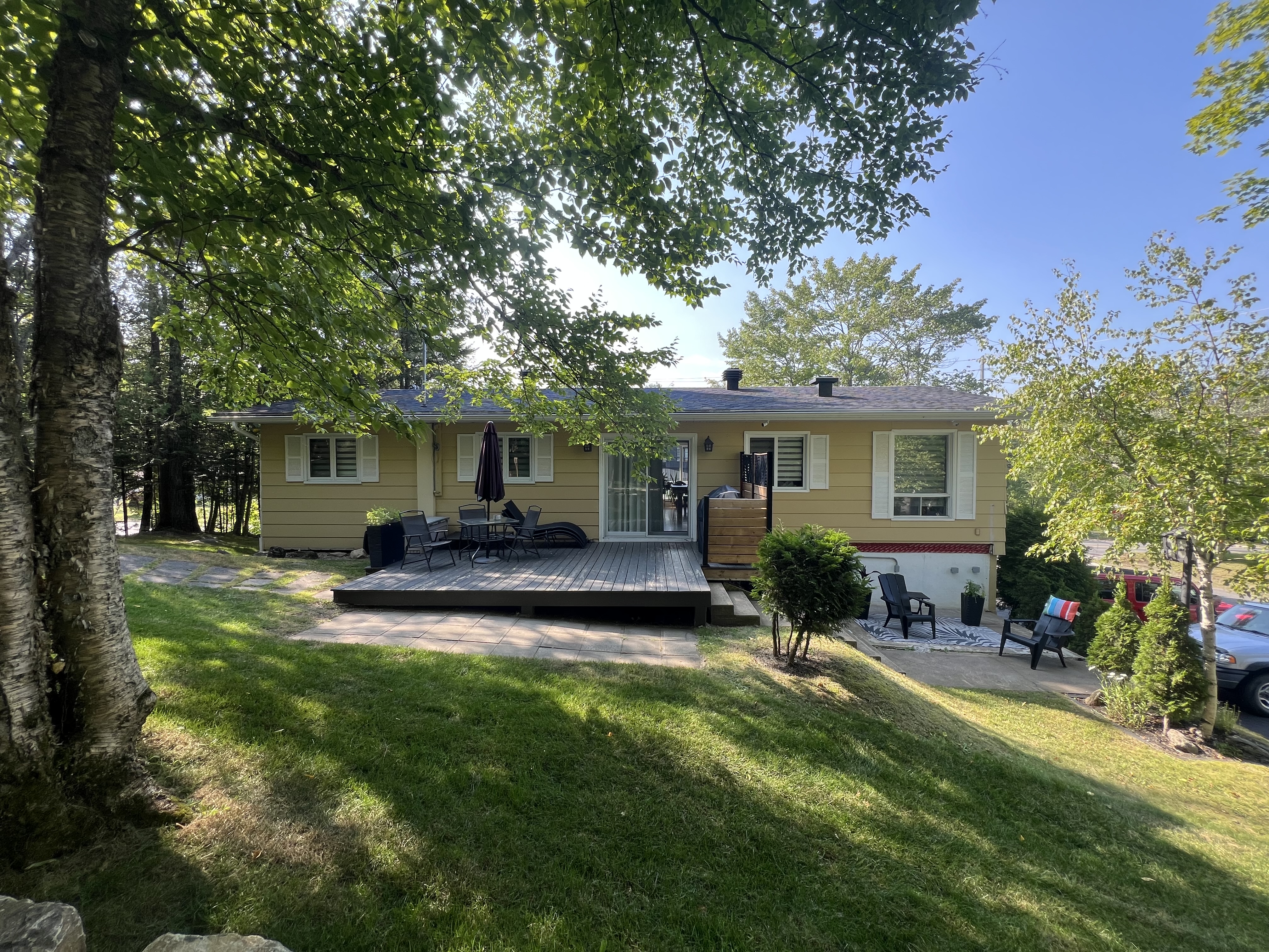 Vue arrière de la maison avec revêtement jaune, patio surélevé en bois et dalles de béton au sol, entourée d'arbres matures.