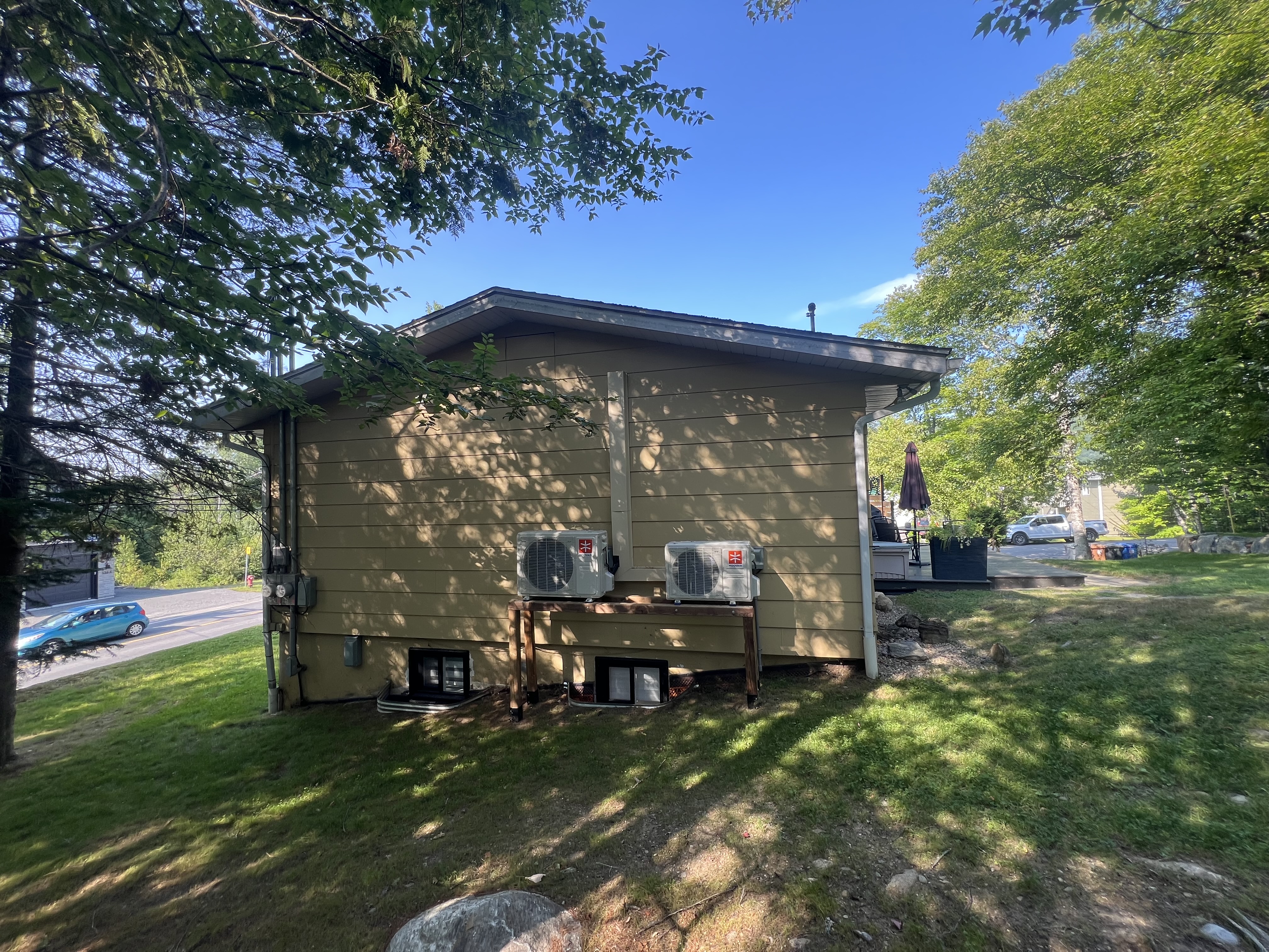 Vue de côté de la maison avec revêtement jaune, montrant deux unités de thermopompe installées sur une structure en bois au-dessus des fenêtres de sous-sol.